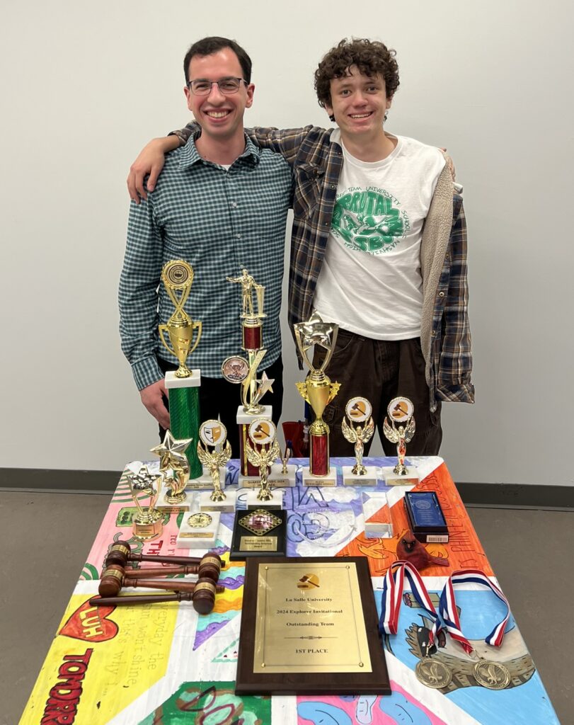 Photo of two team members standing behind a table filled with team trophies.